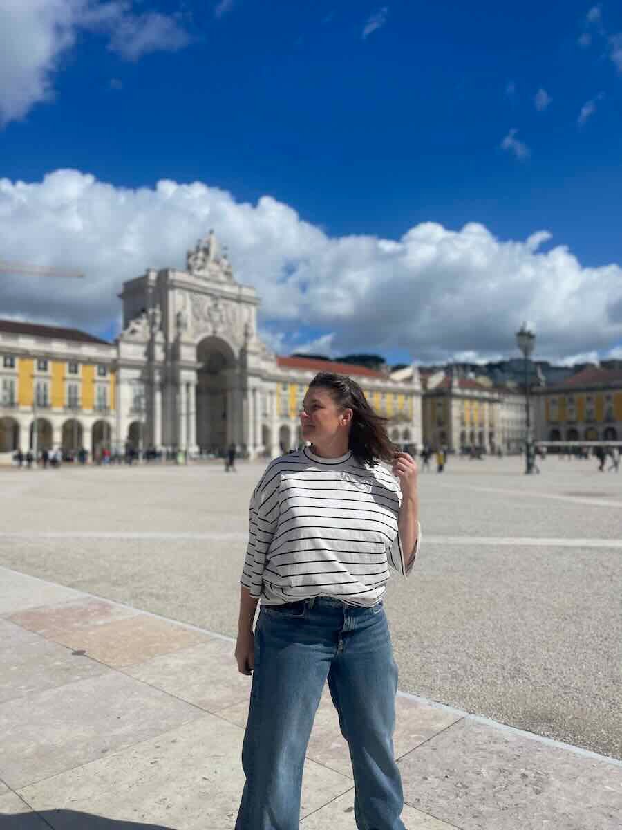 A traveler in casual attire stands confidently in Praça do Comércio, Lisbon's grand square, with the iconic Rua Augusta Arch in the background and a bright blue sky overhead