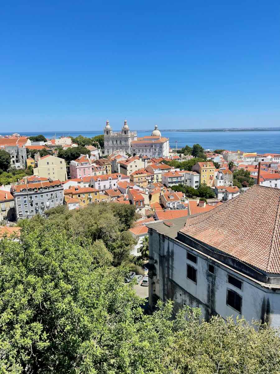 This image captures a stunning view of Lisbon with its iconic red rooftops, the Tagus River in the distance, and the prominent dome of the National Pantheon. The scene is framed by lush greenery under a bright blue sky.