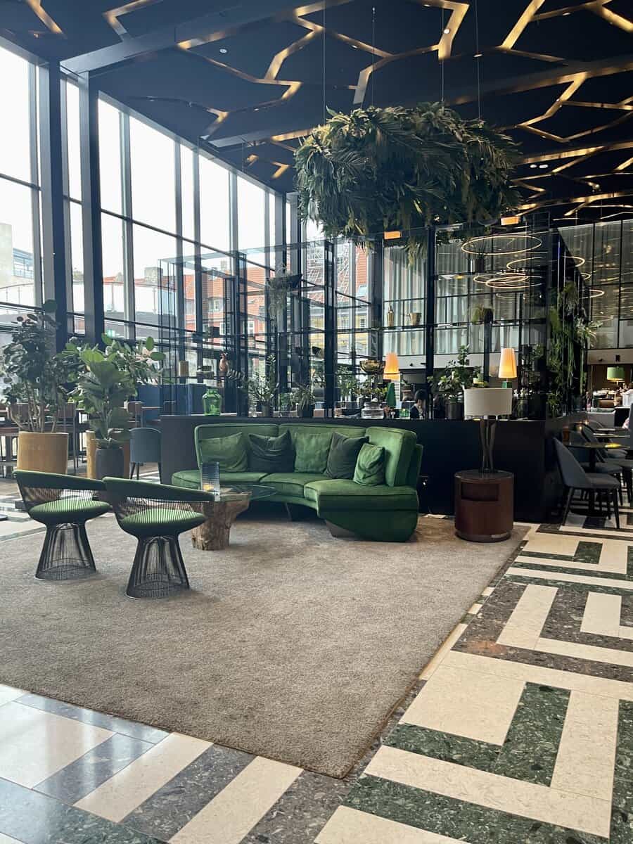 Luxurious lobby area of Hotel Skt Petri featuring floor-to-ceiling windows, a plush green velvet sofa set against a modern decor with abundant greenery and chic lighting, creating a vibrant yet cozy ambiance.