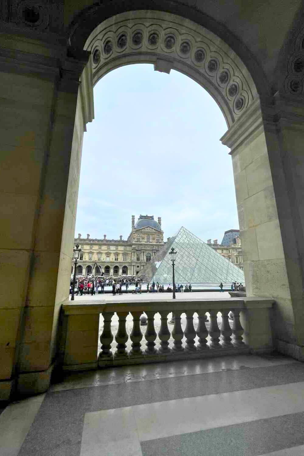 view from inside the Louvre Museum, looking out through an arched doorway towards the iconic glass pyramid and the museum's facade. 