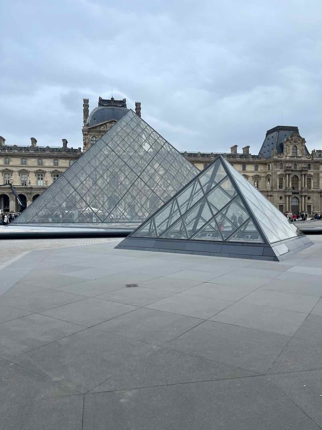 A view of the iconic glass Louvre Pyramid in Paris, with smaller pyramids in the foreground, set against the historic Louvre Palace under a cloudy sky.