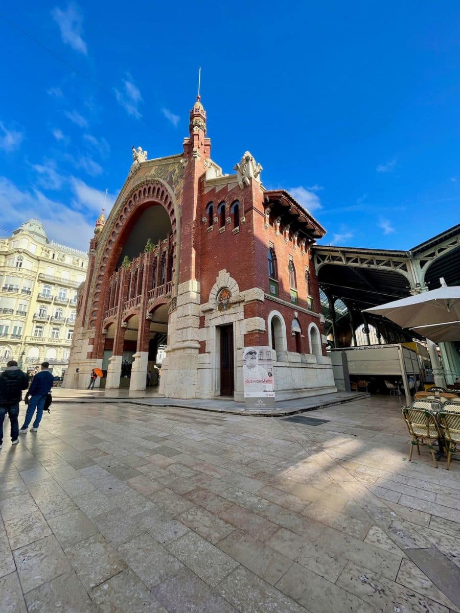Exterior of the Mercado de Colón in Valencia, Spain, showcasing a red-brick facade with an ornate arched entrance, topped with decorative stone carvings and sculptures. The building's striking architectural details are highlighted against a clear blue sky, with a few people and café tables in the foreground.