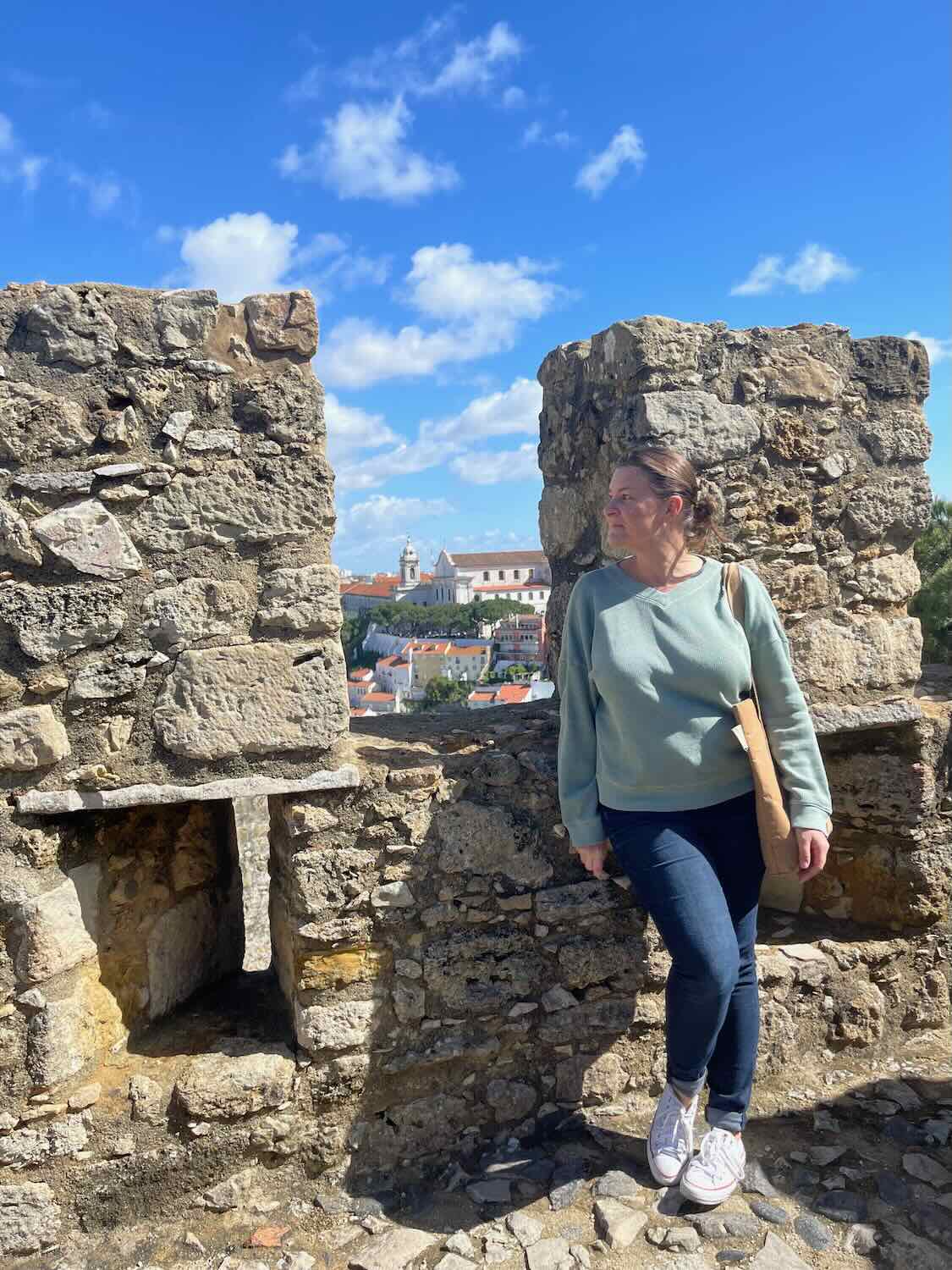 A woman stands contemplatively between ancient stone battlements at a castle in Lisbon, with the cityscape unfurling in the background under a bright blue sky.