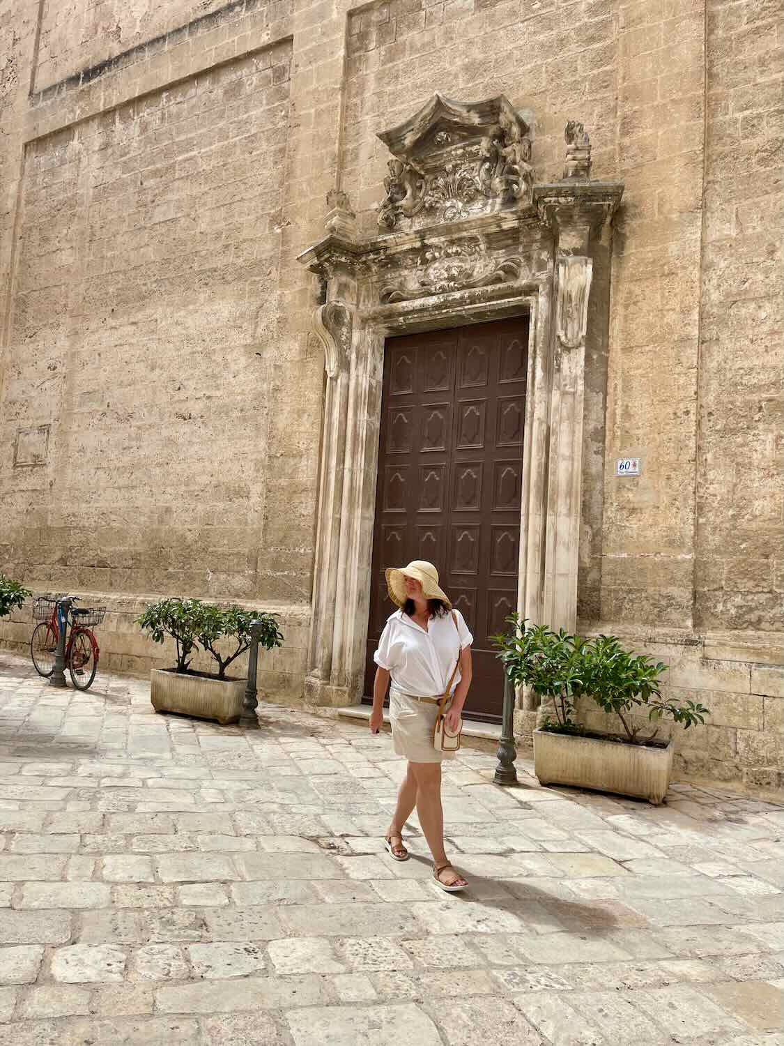 A woman wearing a white blouse, beige shorts, sandals, and a wide-brimmed straw hat walks along a cobblestone street in Monopoli, Italy. She is positioned in front of an ornate wooden door set within a grand, historic stone building.