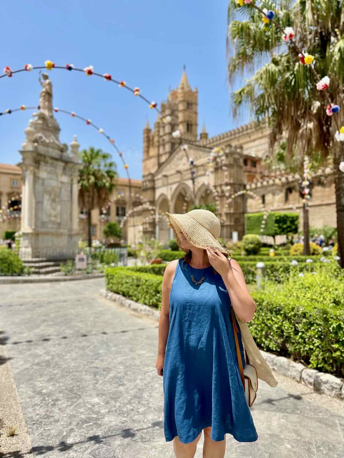 A woman in a blue dress and straw hat stands in a sunny courtyard in Palermo, Sicily, with historic buildings and a statue in the background. Colorful decorations hang across the courtyard, adding a festive atmosphere.