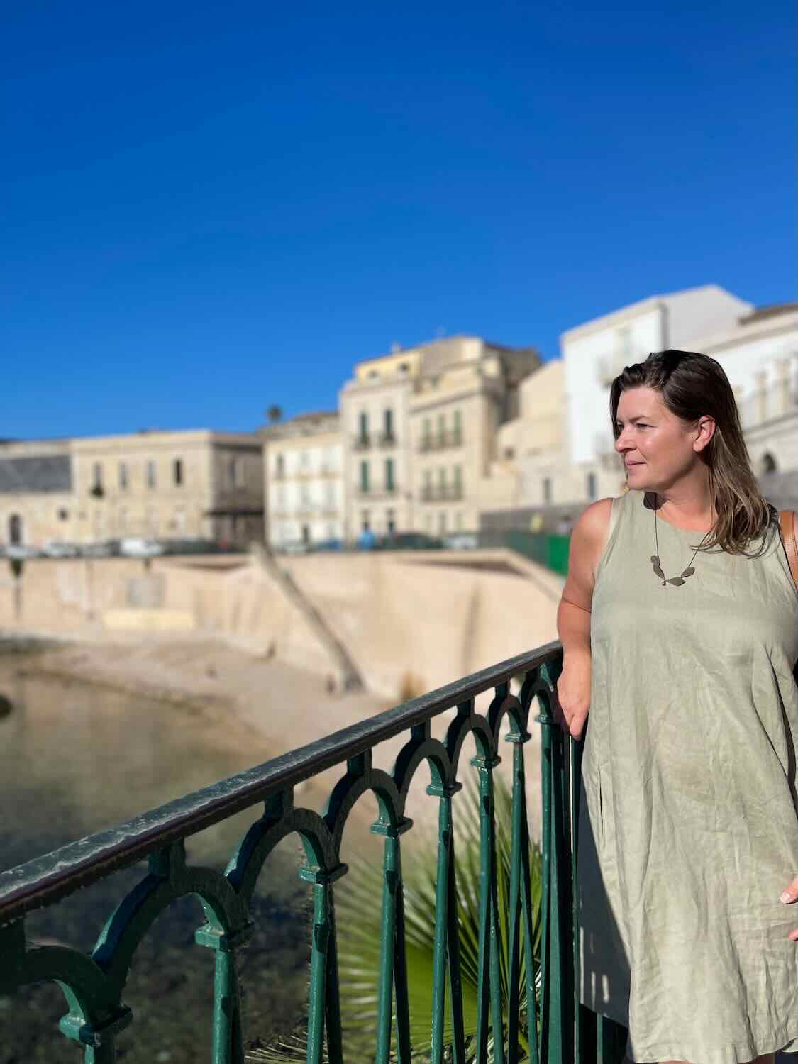 A woman in a light green dress stands on a railing overlooking the waterfront in Siracusa, Sicily. The historic buildings and clear blue sky in the background add to the picturesque scene.