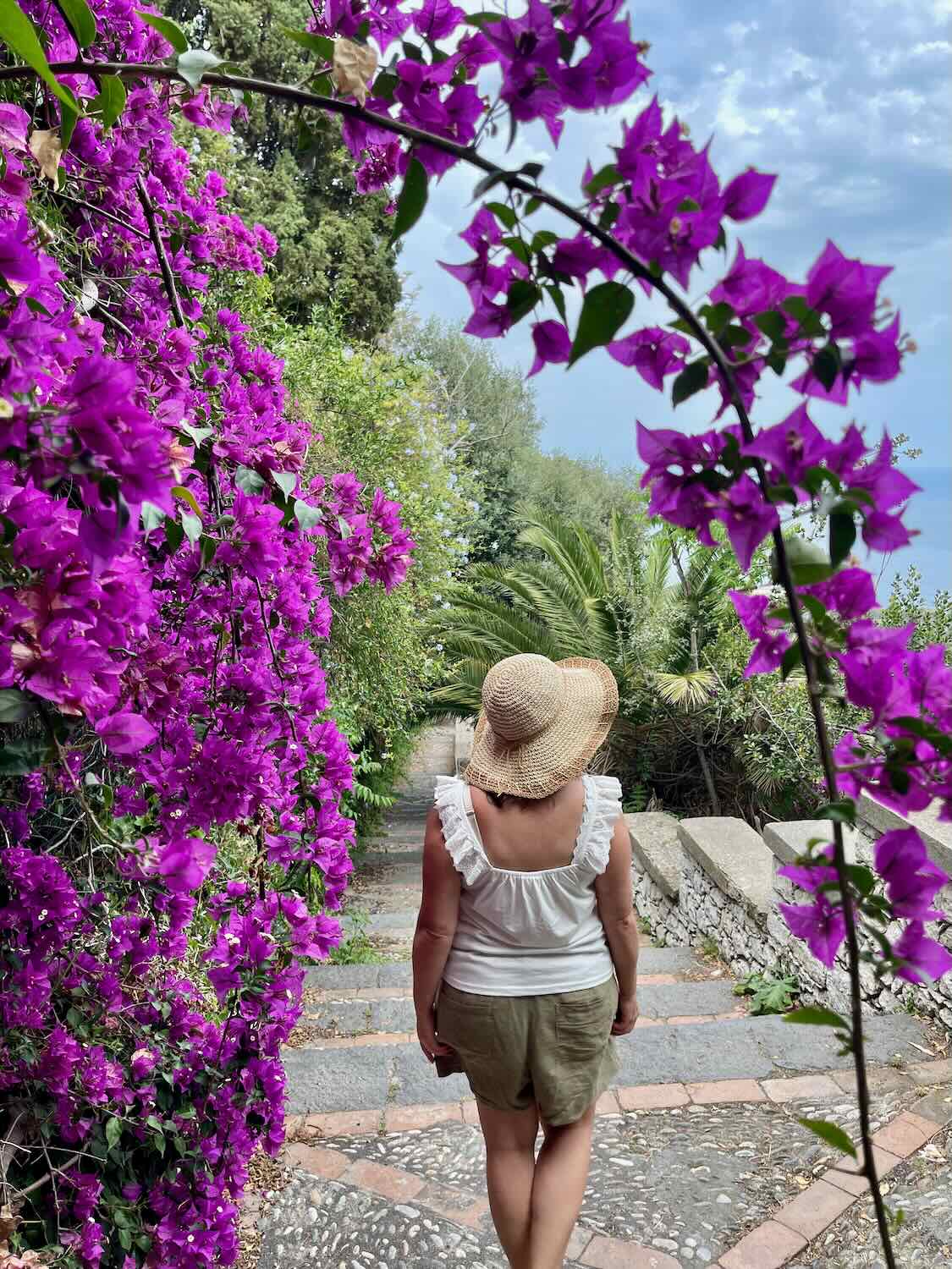 A woman in a straw hat and light summer outfit walks through a lush garden archway adorned with vibrant purple bougainvillea flowers, adding a touch of natural beauty to the serene setting.