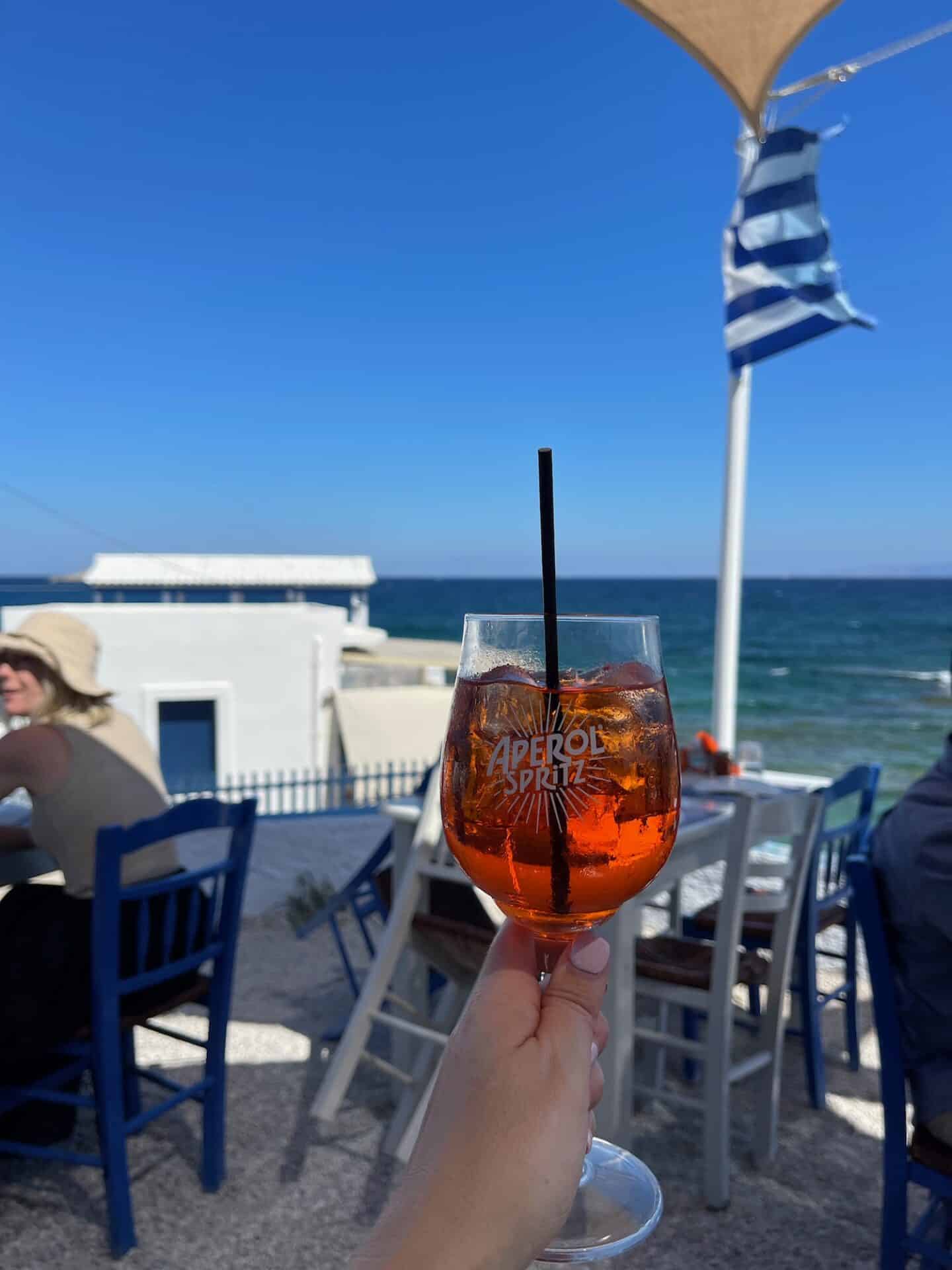 Close-up of a hand holding a cocktail with a blue and white striped Greek flag fluttering in the background at a seaside café.