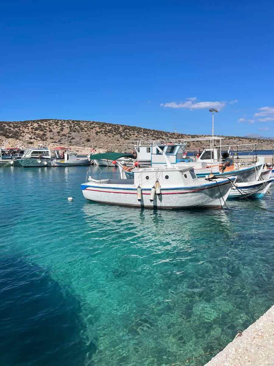 This image shows several small boats docked in a serene harbor on Milos Island, Greece. The water is a vibrant, clear turquoise, and the boats are lined up along the dock. In the background, low, rocky hills stretch under a bright blue sky.