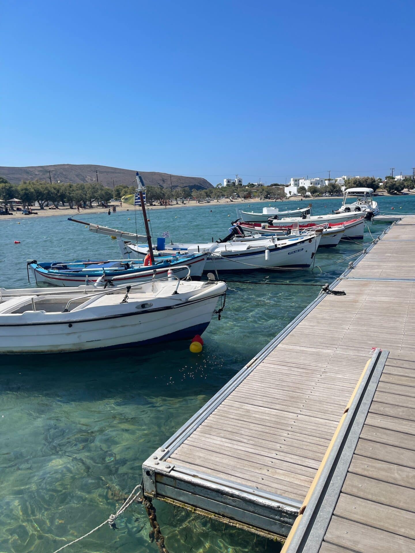 A wooden dock extends into a calm bay with several boats moored along it, set against a clear blue sky.
