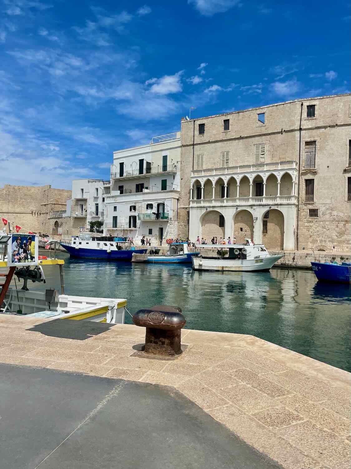 This image captures a picturesque harbor scene in a historic coastal town. The water is calm, with small fishing boats moored along the edges. Surrounding the harbor are aged stone buildings, including one with beautiful arches and balconies, suggesting a blend of historic and Mediterranean architecture. The bright blue sky enhances the vibrancy of the scene, while the stone pier in the foreground gives a sense of peaceful daily life by the water. This setting seems perfect for a relaxing stroll, likely in a coastal town in Southern Europe.