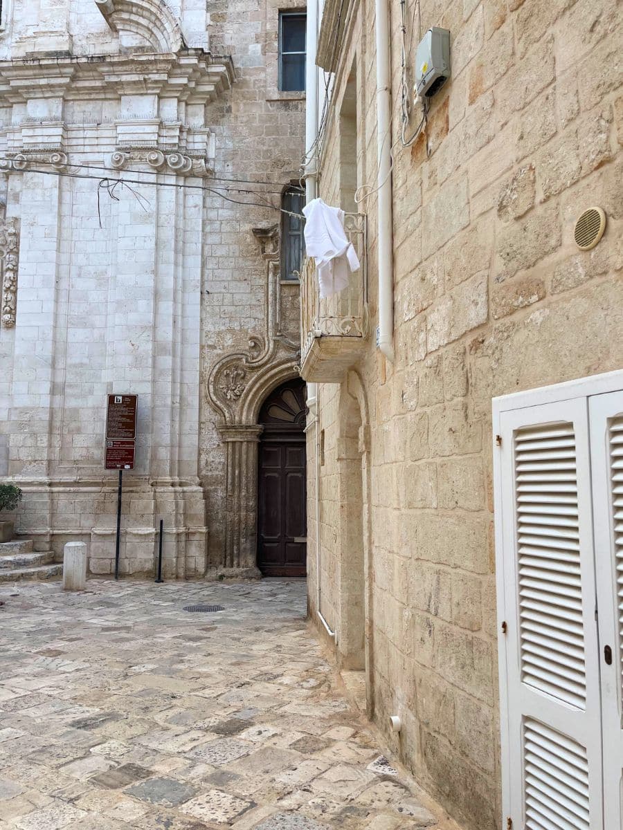 A quiet stone-paved alley in Monopoli, Italy, featuring the ornate façade of a historic church, a wooden door, and a small balcony with white laundry hanging.
