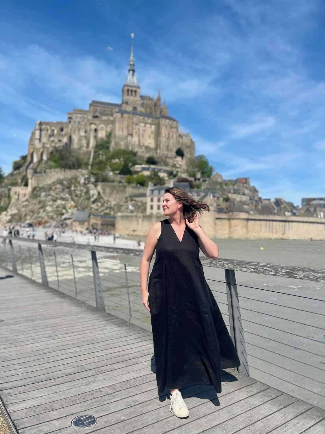 Solo female traveler standing on a wooden boardwalk with Mont Saint Michel in the background, highlighting the personal experience of visiting.
