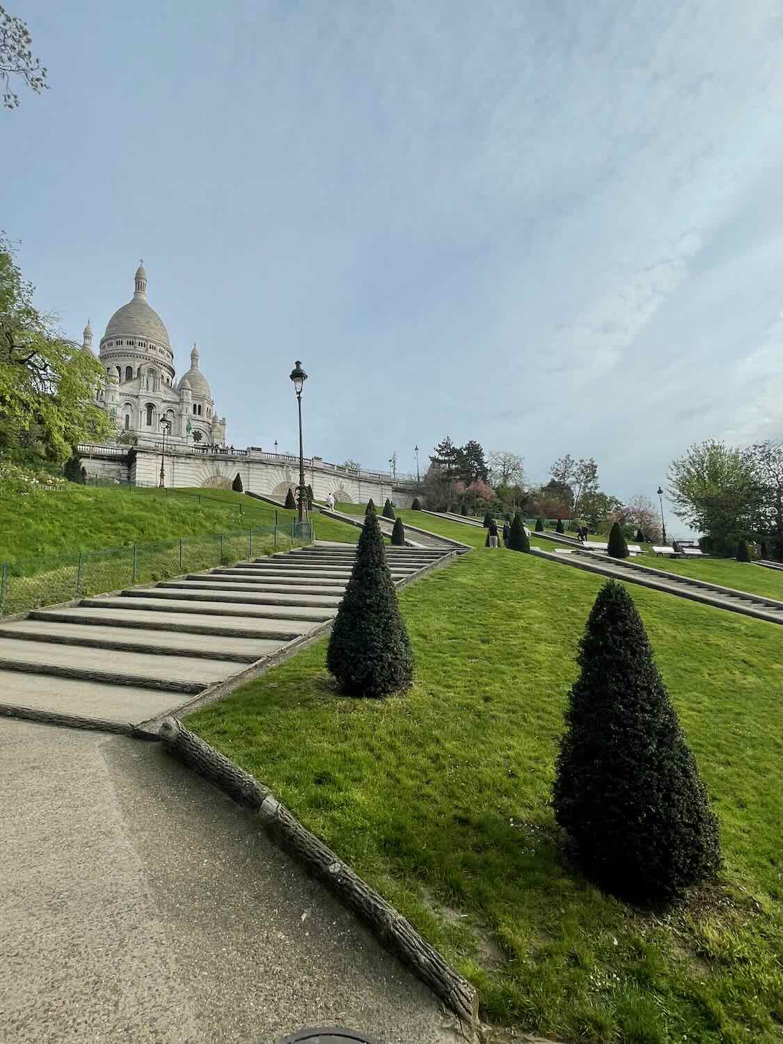 View of the Sacré-Cœur Basilica from the bottom of a staircase leading up the Montmartre hill, showcasing the lush greenery and clear skies.