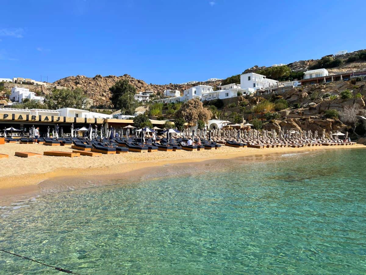 A tranquil beach scene showing clear turquoise waters and a sandy shore. There are rows of sunbeds with umbrellas