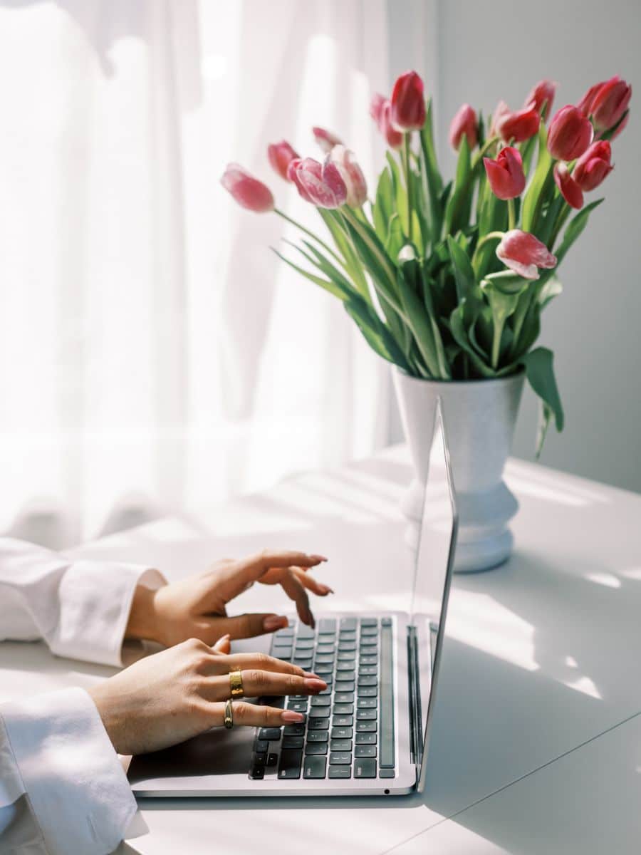 Woman using laptop at a bright, feminine workspace with pink tulips, representing productivity with a Notion travel template for women.