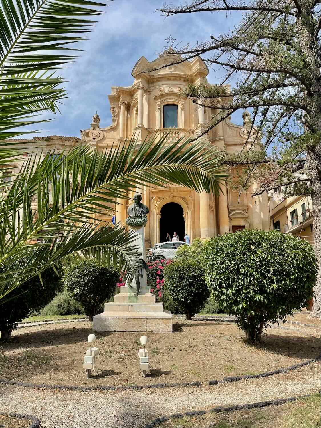 Baroque church in Noto, Sicily, framed by palm fronds and greenery, with a statue in the foreground."

Let me know if you'd like any refinements.