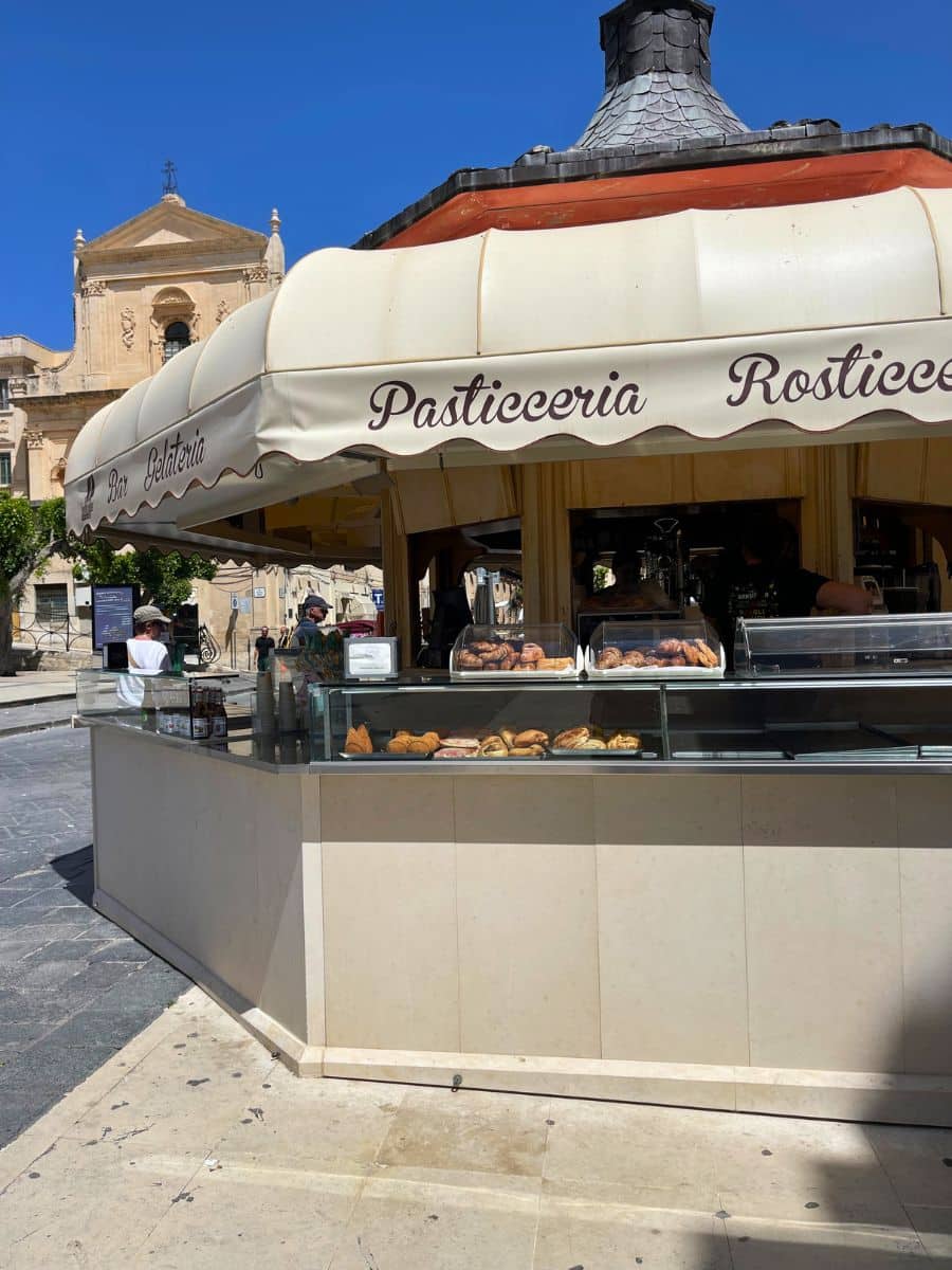 A quaint outdoor pastry and snack stand in Noto, Sicily, with a cream-colored canopy labeled 'Pasticceria', 'Rosticceria', 'Bar', and 'Gelateria'. The display case is filled with assorted pastries, croissants, and savory items. Behind the stand, a historic Baroque church with ornate architectural details is visible, set against a clear blue sky.