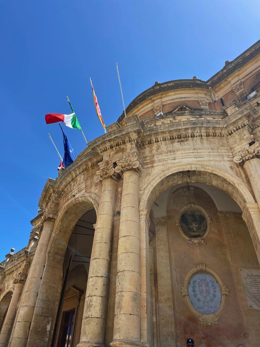 An impressive Baroque-style building in Noto, Sicily, with grand arched columns and detailed stone carvings. Multiple flags, including the Italian and European Union flags, wave above the entrance. Decorative frescoes and emblems adorn the wall beneath the arches, and the clear blue sky highlights the structure’s architectural elegance.