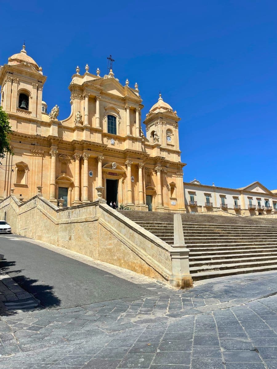 The grand façade of Noto Cathedral in Sicily, showcasing stunning Baroque architecture with tall columns, ornate sculptures, and twin bell towers. A wide stone staircase leads up to the entrance, with the cathedral glowing warmly under a clear blue sky. Adjacent buildings with arched windows line the background.