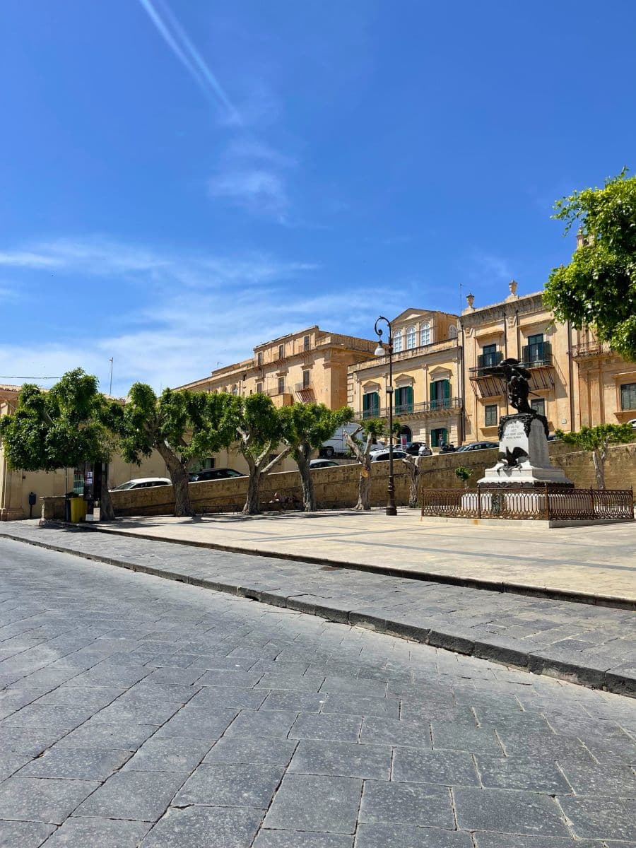 A charming town square in Noto, Sicily, featuring a small statue monument surrounded by a decorative iron fence. Neatly pruned trees line the stone-paved area, casting dappled shade. In the background, elegant historic buildings with balconies and green shutters stand under a vibrant blue sky.