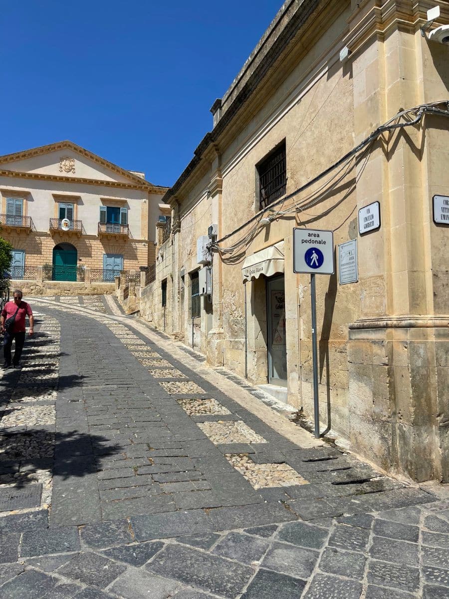 A charming sloped pedestrian street in Noto, Sicily, paved with a mix of dark stone tiles and pebbles. On the right, rustic stone buildings line the walkway, including a shop with a canopy. A man walks uphill, and at the top stands a grand neoclassical building with a triangular pediment and green doors, framed by a clear blue sky.