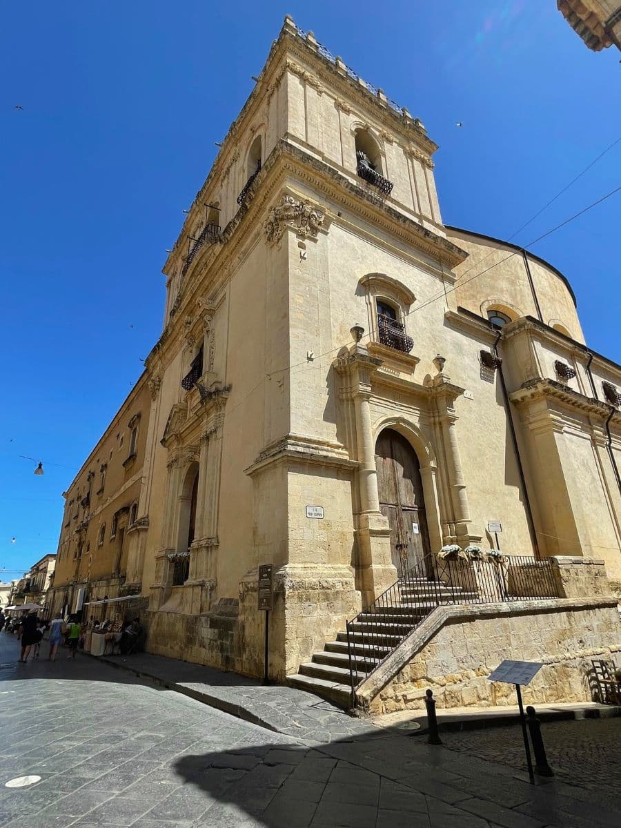 A tall Baroque church building in Noto, Sicily, with a prominent bell tower and ornate architectural details. The façade features arched windows, balconies with wrought-iron railings, and a grand wooden door accessed by a flight of stone stairs. The church stands at a street corner, casting shadows on the cobbled street below, while a few pedestrians stroll nearby under a clear blue sky.