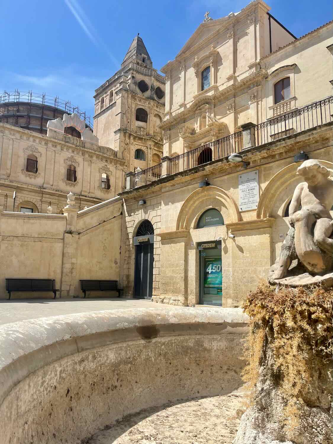 Baroque building in Noto, Sicily, with a bell tower and arched windows. A small fountain with a weathered statue sits in the foreground.