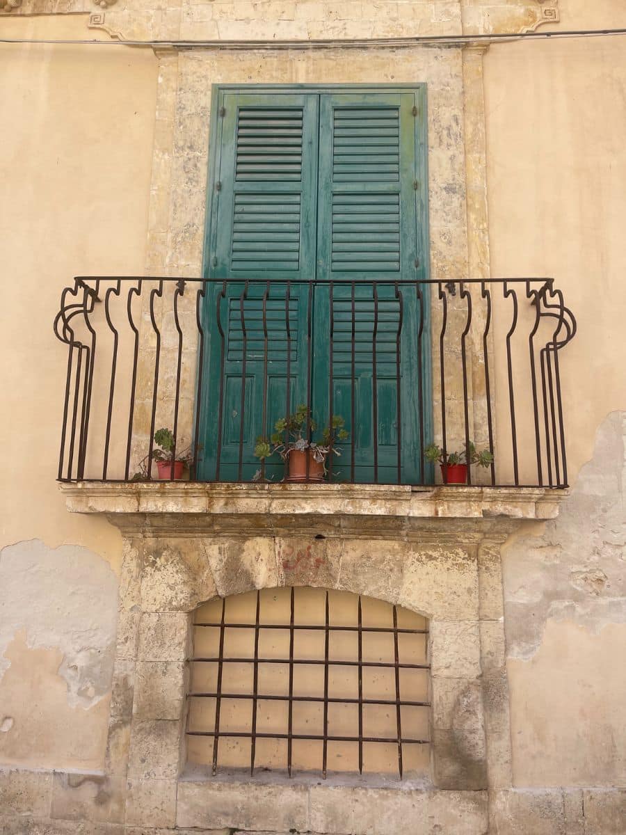 A charming balcony in Noto, Sicily, featuring a pair of green wooden shutter doors framed by aged limestone. The wrought iron railing curves gently outward, adorned with a few small potted plants. The wall has a warm, weathered patina with visible patches of peeling plaster. Below the balcony, there is a barred arched window set in rustic stone, contributing to the historic and authentic character of the building façade.
