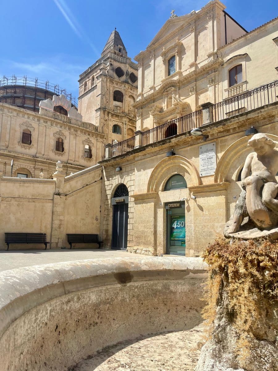 "A sunlit square in Noto, Sicily, showcasing the ornate Baroque facade of a historic church with detailed stonework and a bell tower in the background. Below the church, arched doorways house a bank and an entrance marked 'CREDEN'. In the foreground, a weathered stone fountain features a sculpted figure partially covered in moss, and a curved basin. Benches line the perimeter of the square, inviting passersby to rest and take in the view under the bright Sicilian sky.