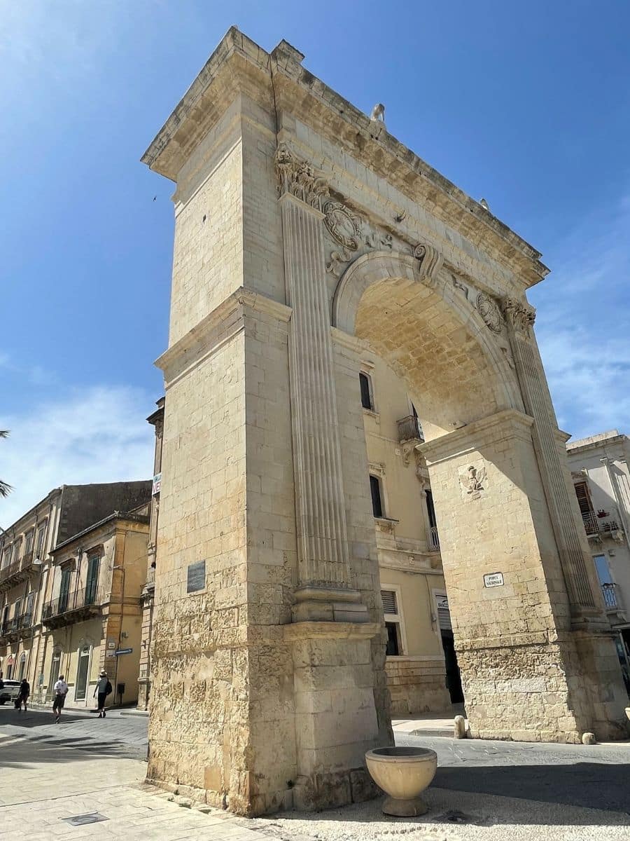 A grand stone archway in Noto, Sicily, known as the Porta Reale, stands tall against a clear blue sky. The neoclassical structure features Corinthian columns, ornate carvings, and sculptural details at the top, including a crowned emblem and decorative flourishes. The arch leads into a street lined with traditional Sicilian buildings with balconies and shuttered windows. A few pedestrians stroll through the sunlit square beneath the arch, emphasizing the monument's scale and historical charm.