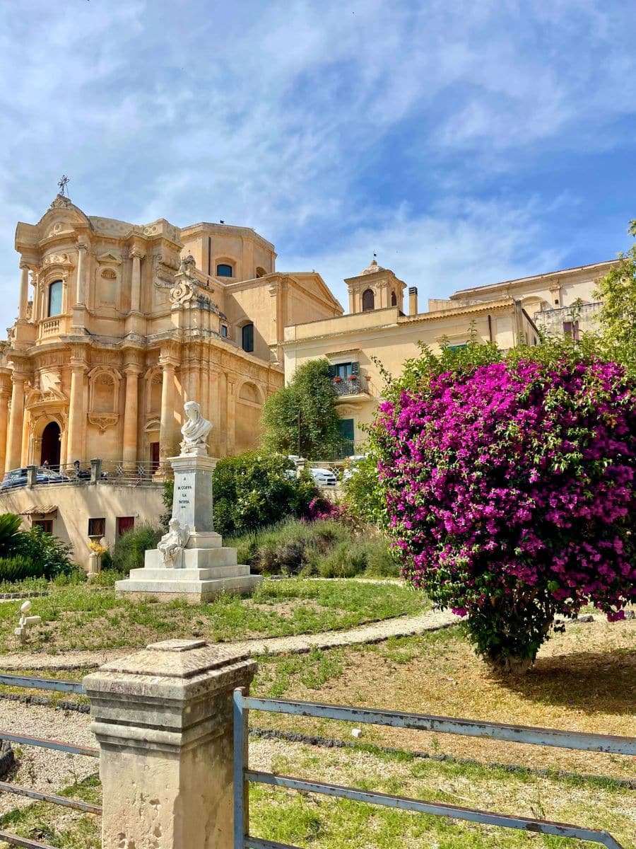 A picturesque garden in Noto, Sicily, featuring a white marble statue on a pedestal in the center, surrounded by greenery and flowering bushes. To the right, a vibrant bougainvillea bush blooms with bright magenta flowers. In the background stands an elegant Baroque church with ornate architecture, including arched windows, decorative columns, and sculptural details. The golden-hued building contrasts beautifully with the bright blue sky and lush garden, creating a serene and scenic view.