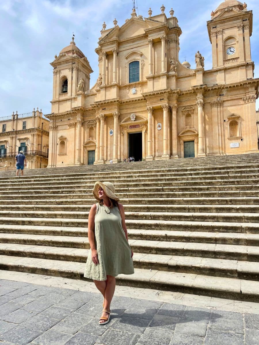 Melissa wearing a light green sleeveless dress and a wide-brimmed straw hat stands on the stone steps of the grand Noto Cathedral in Sicily, Italy. The stunning Baroque-style cathedral towers in the background with its detailed architecture, twin bell towers, and ornate columns. The woman poses casually, looking to the side and smiling, basking in the sunlight. The light golden hue of the cathedral contrasts beautifully with the blue sky and adds to the charm of the historic setting.