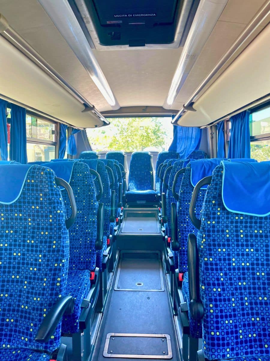 Interior of a modern intercity bus with rows of blue patterned seats featuring armrests and headrest covers. The aisle leads towards the back of the bus, with overhead compartments and curtains on the windows. Natural light streams in through the windows, complementing the bright and clean interior.