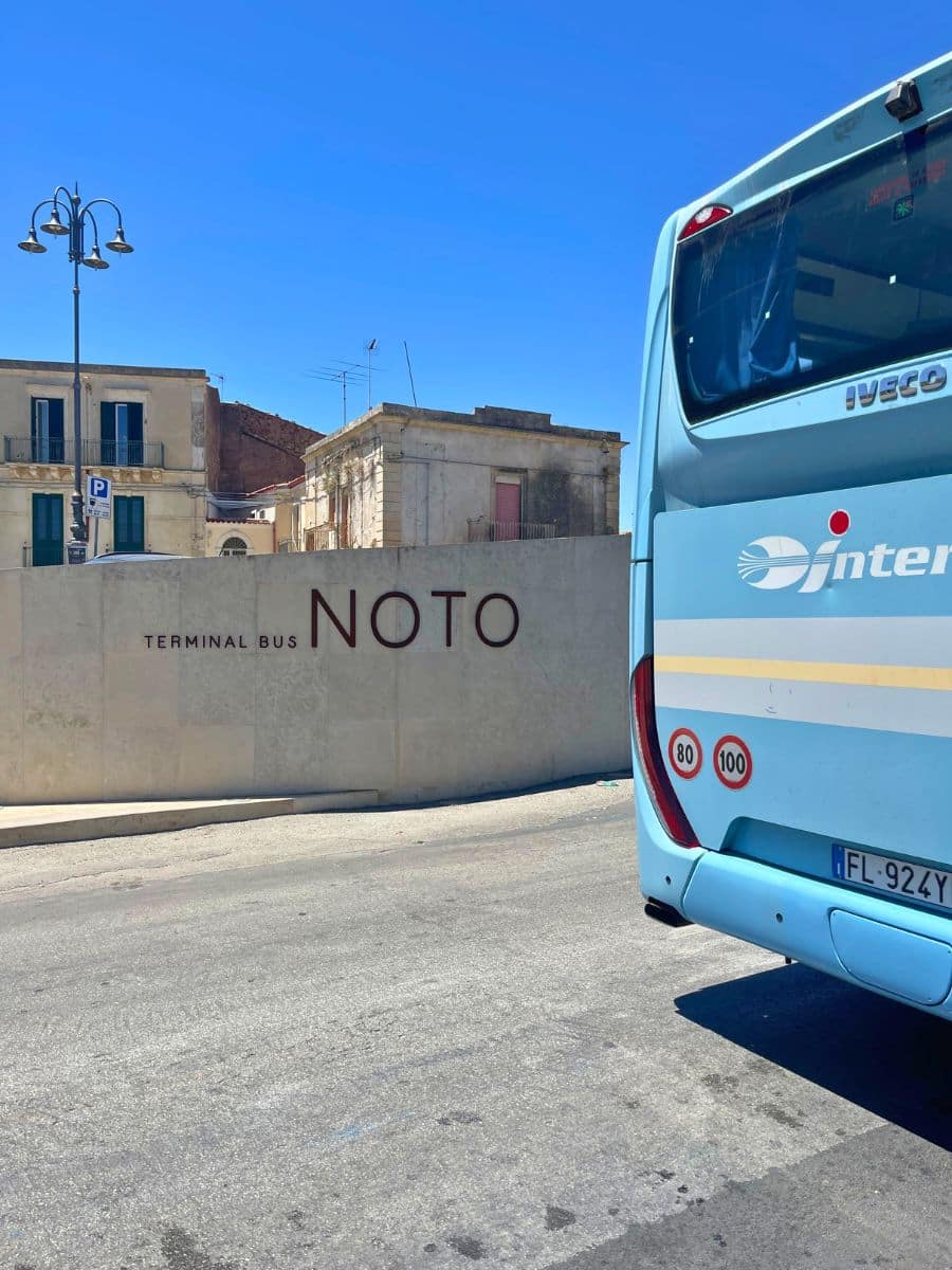 A sunny day at the Noto bus terminal, marked by a modern beige stone wall with 'NOTO' inscribed in bold, elegant letters. A blue intercity bus is parked beside it, partially visible in the foreground. In the background, classic Italian buildings with green shutters and balconies showcase the town’s historic charm, set against a vivid blue sky.