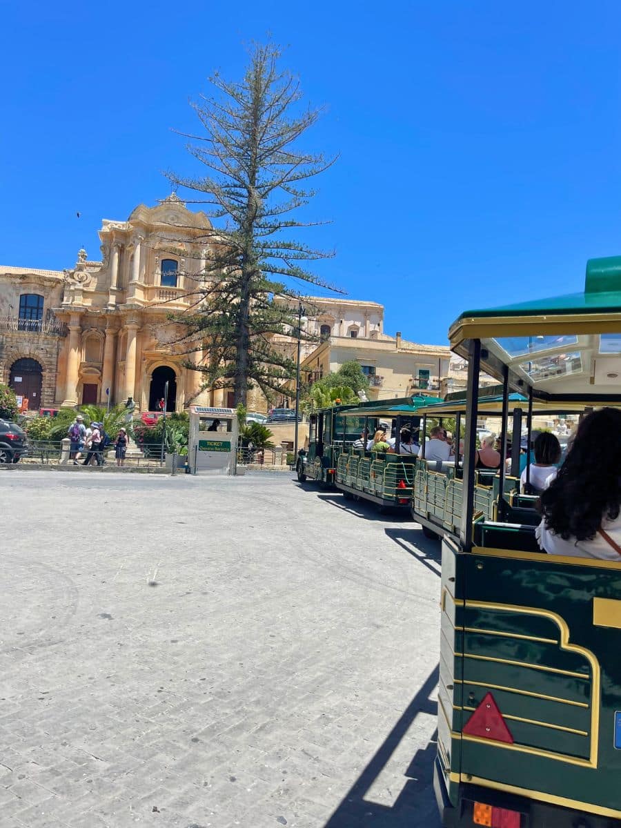 A scenic view of a Baroque-style church in Noto, Sicily, Italy, with its ornate façade featuring arched entrances, columns, and sculptural details. In the foreground, a green and yellow tourist train with open carriages is filled with passengers enjoying a city tour. A tall, sparse tree stands prominently in front of the church, adding contrast to the architectural backdrop. The sky is bright blue, and people can be seen walking near a ticket booth, contributing to a lively, sunny atmosphere in the town square.