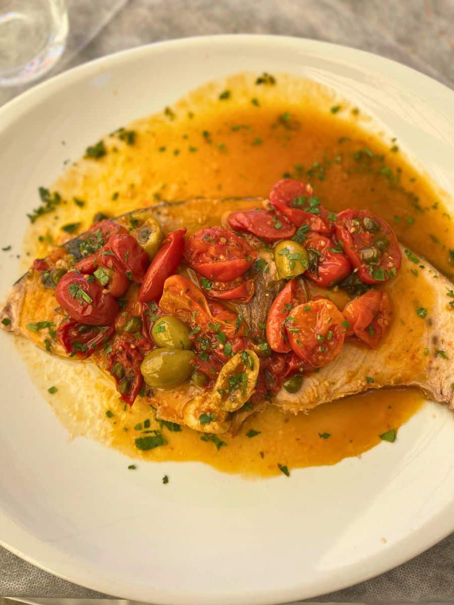 A beautifully plated Mediterranean-style fish dish, served with a flavorful sauce of cherry tomatoes, green olives, capers, and fresh herbs. The whole fish fillet lies in a vibrant orange-red broth on a white plate, garnished with chopped parsley. The dish captures the essence of Sicilian cuisine, combining fresh seafood with bold, sun-ripened ingredients.