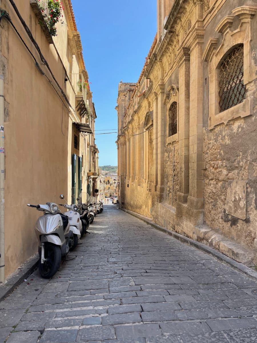A charming narrow cobblestone alley in Noto, Sicily, lined with parked scooters on one side. The buildings feature warm-toned stucco and intricate Baroque architectural details, including arched windows and stone carvings. Flower pots hang from balconies above, adding a touch of color. The alley gently slopes downwards toward a sunlit square in the distance.