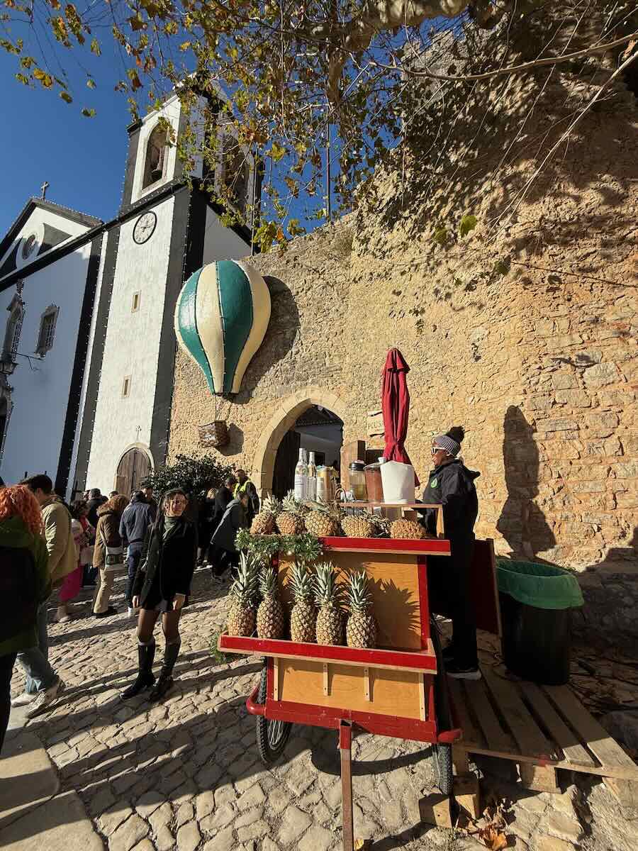 A festive outdoor scene in Obidos with a pineapple cart under a large decorative hot air balloon next to an old stone wall and church, surrounded by visitors enjoying the event.