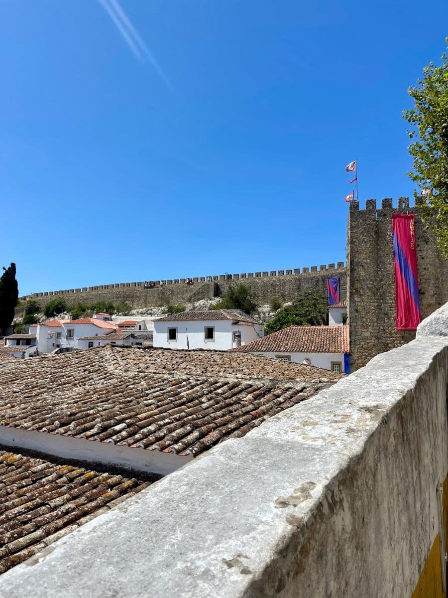 View of the medieval castle walls in Óbidos, Portugal, stretching across the background under a bright blue sky, with red-tiled rooftops of whitewashed houses in the foreground and colorful banners hanging from the stone tower.