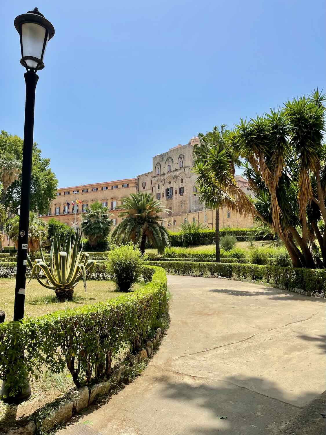 A lush garden path lined with trimmed hedges and palm trees leads to the historic Palazzo dei Normanni in Palermo, Sicily. The building's ornate architecture is visible under a clear blue sky.