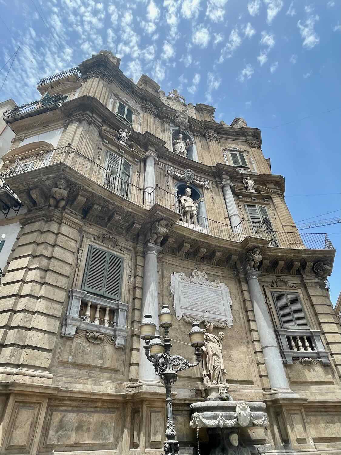 The intricate façade of Quattro Canti, a historic Baroque square in Palermo, Sicily, featuring statues and detailed carvings. The building's weathered stonework stands out against a partly cloudy sky.