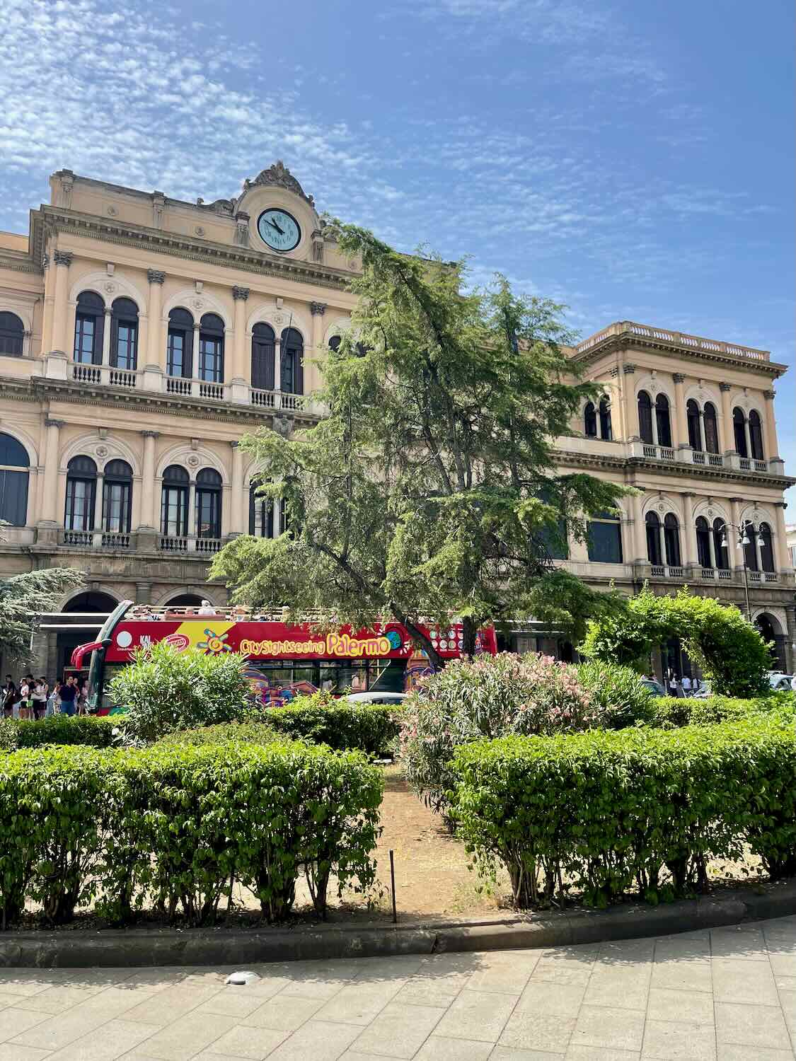 A historic building in Palermo, Sicily, featuring arched windows and a clock at the top. A red sightseeing bus labeled 'City Sightseeing Palermo' is parked in front of the building, with lush green bushes and trees in the foreground under a partly cloudy sky.