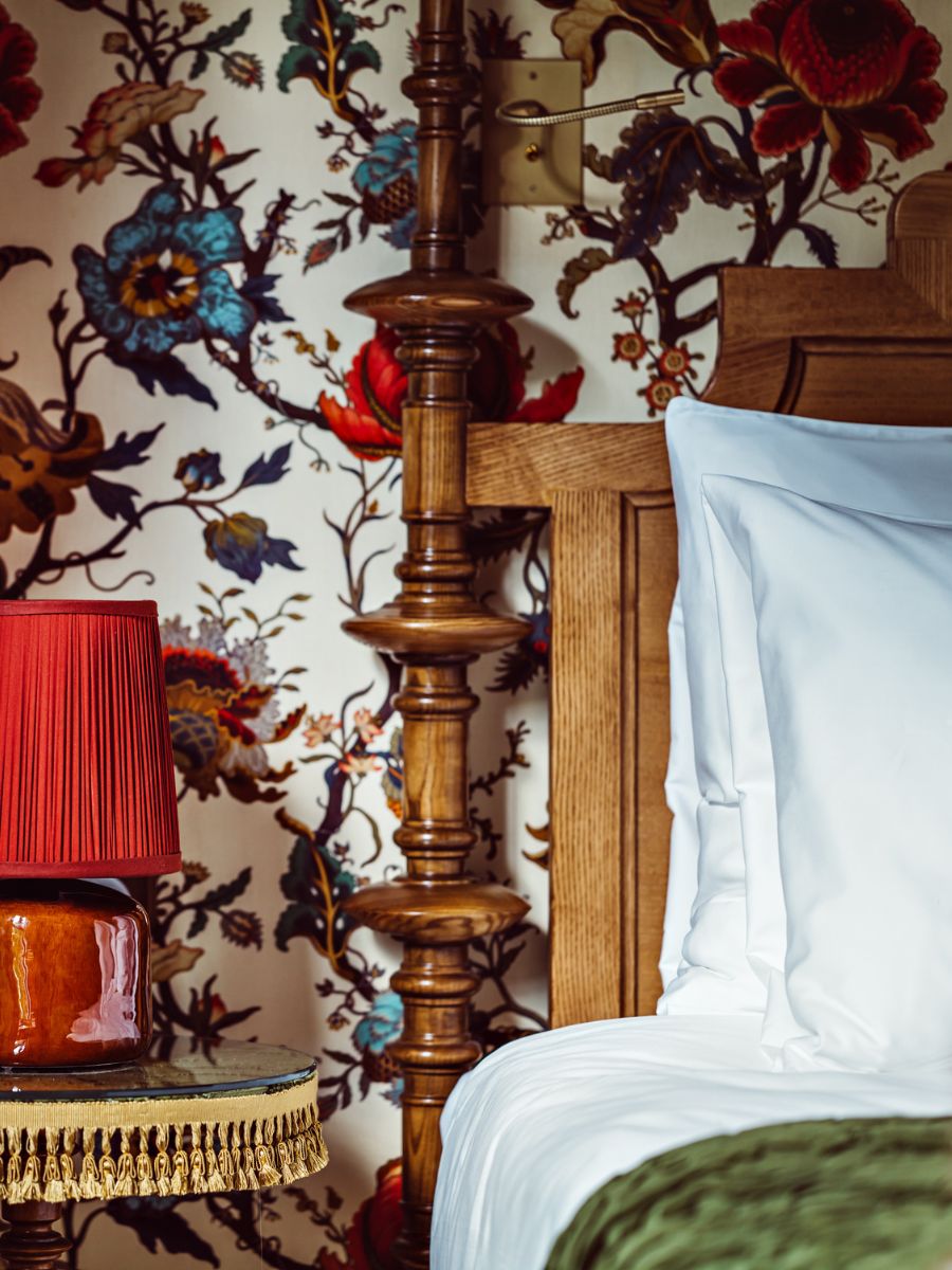Cozy bedroom corner with a carved wooden bedframe, crisp white linens, a red pleated lamp on a vintage nightstand, and vibrant floral wallpaper in the background.