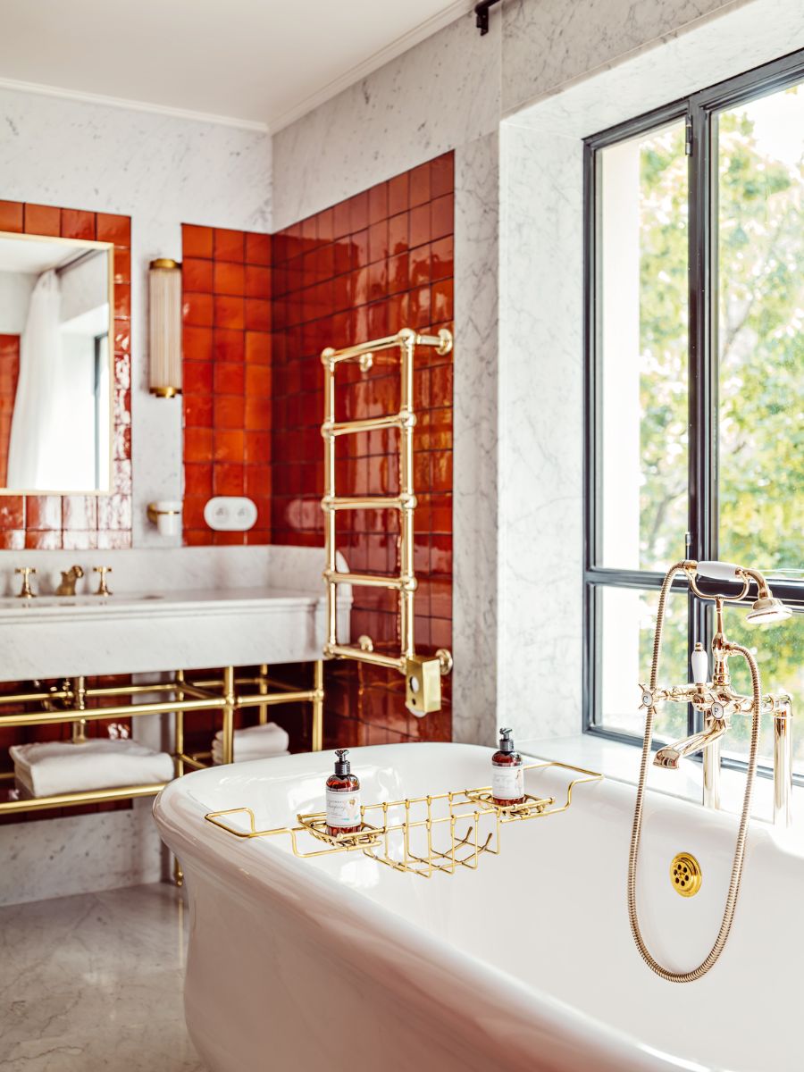 Elegant bathroom with a freestanding white tub, gold fixtures, red tile accents, marble surfaces, and large windows bringing in natural light.