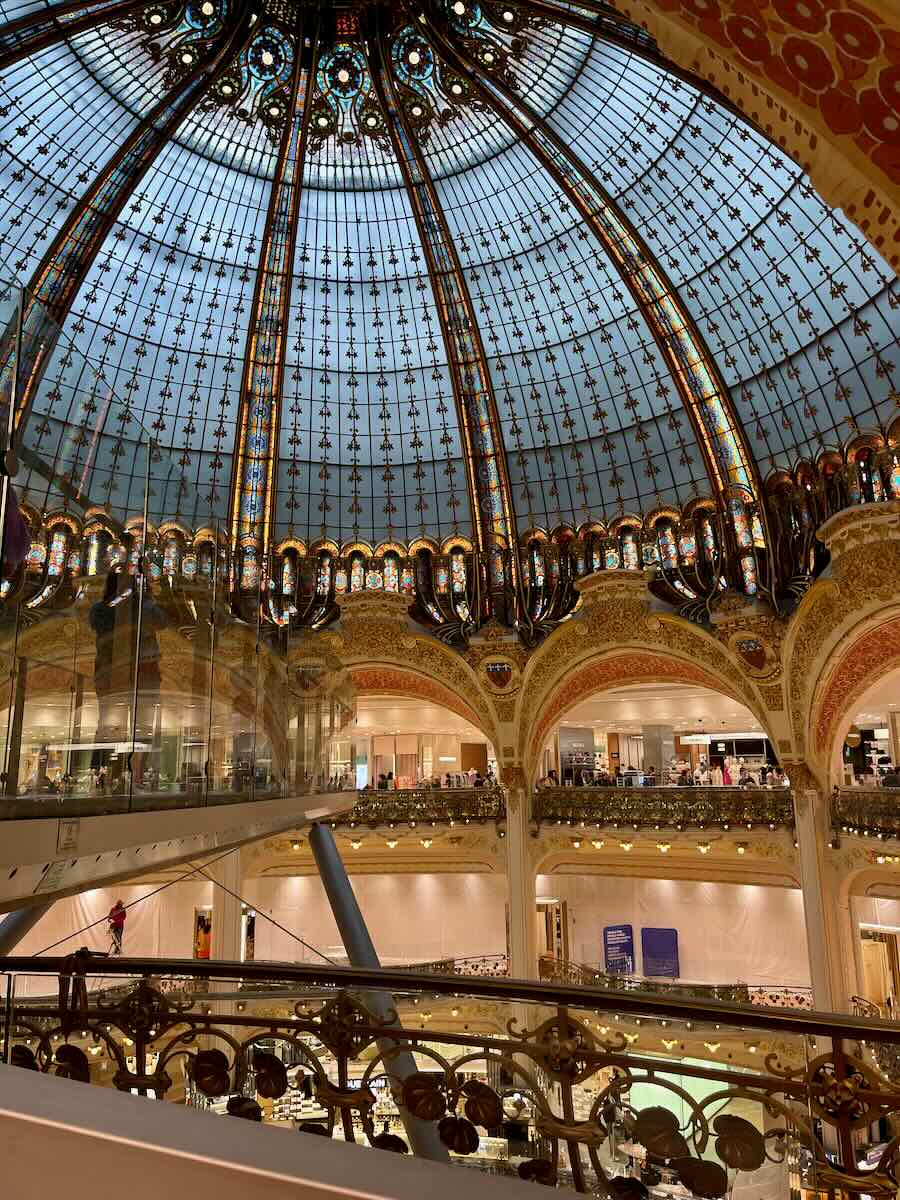 Interior view of a grand dome in Paris with intricate stained glass, ornate gold detailing, and elegant arches in a multi-level shopping space.