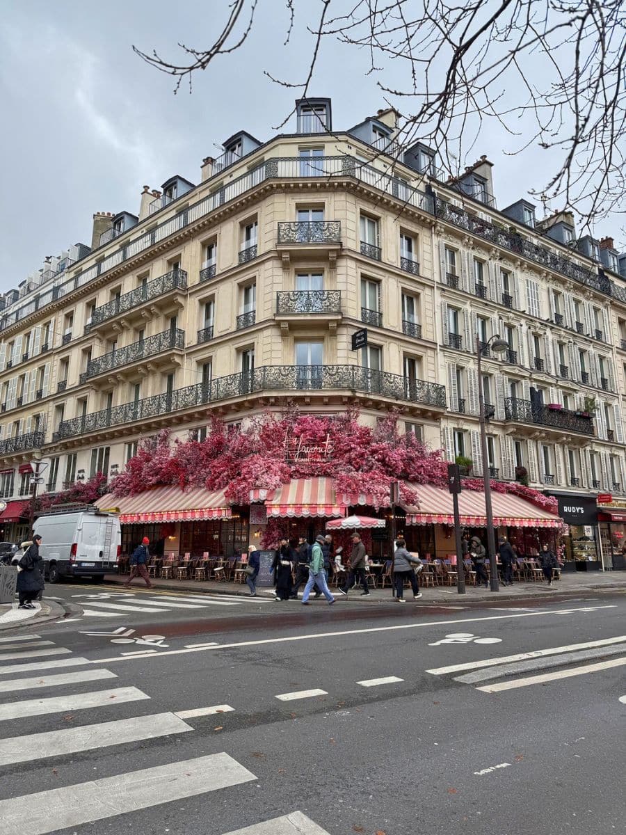 A Parisian building with ornate balconies, featuring a café adorned with pink flowers and striped awnings at the corner of a busy street.