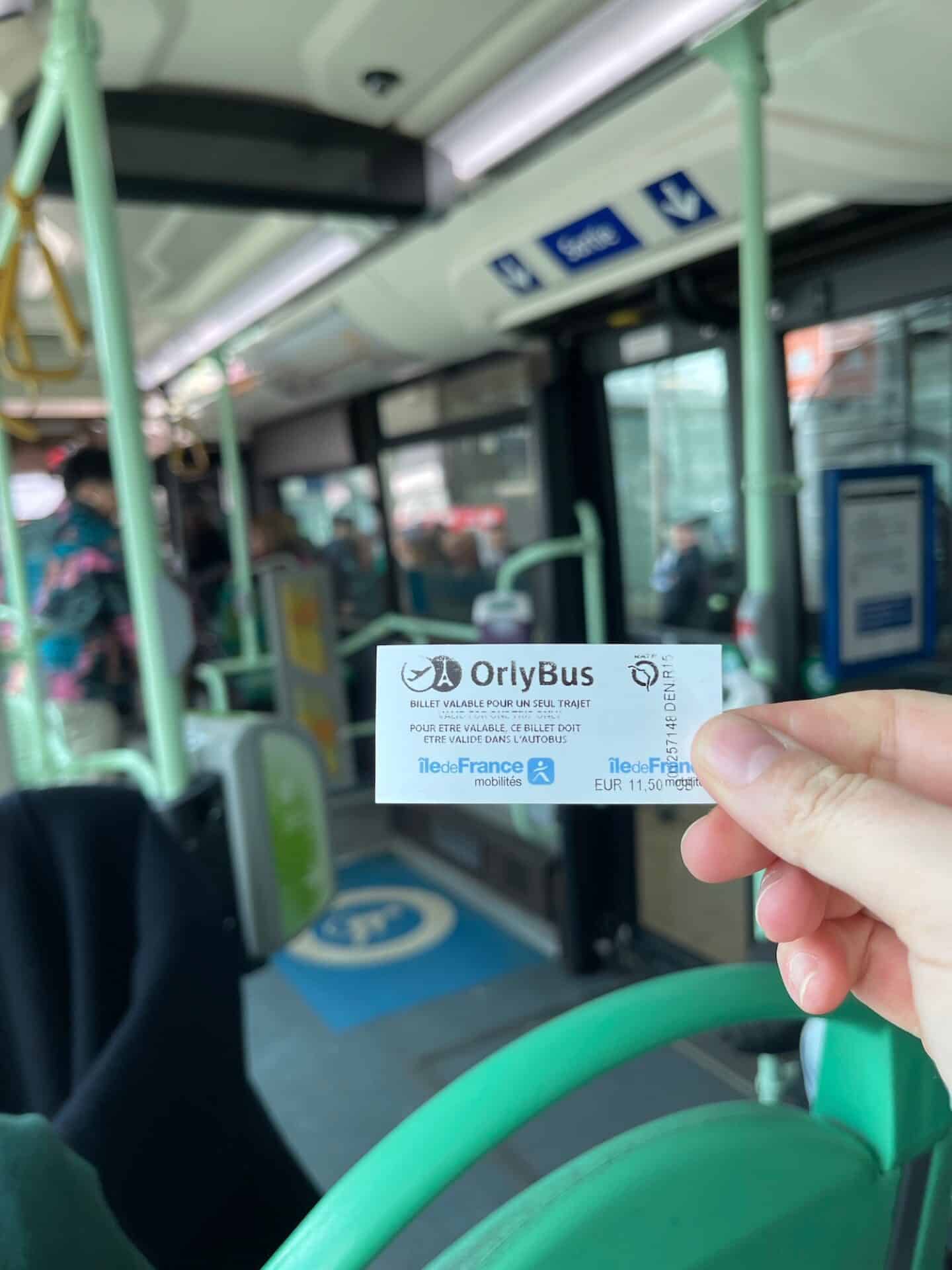 A hand holding an OrlyBus ticket inside the bus, showing the fare of EUR 11.50. The background features the interior of the bus with passengers and a 'Sortie' (exit) sign.