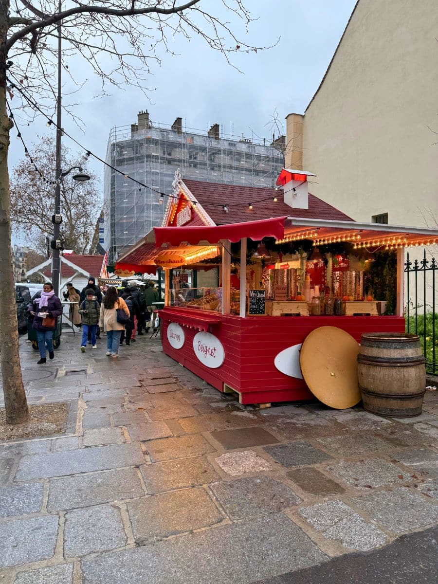 A charming red Christmas market stall in Paris offering treats like churros and beignets. The stall is decorated with warm string lights and holiday garlands. Nearby, people stroll on cobblestone paths under bare trees on a cloudy winter day. A wooden barrel and table are placed beside the stall.