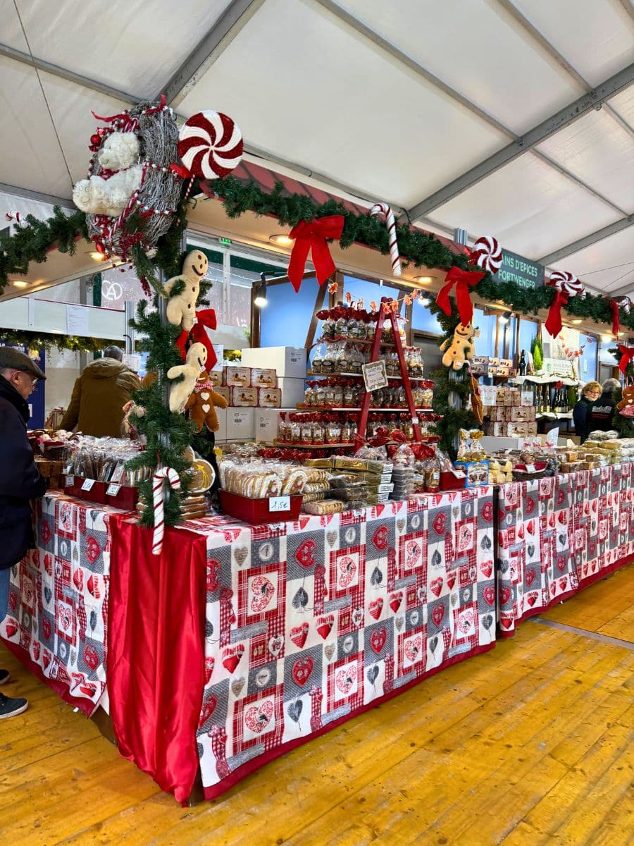 A festive stall at a Paris Christmas market, decorated with garlands, red bows, candy canes, and gingerbread figures. The table is covered with a holiday-themed cloth, displaying an assortment of baked goods and treats, including cookies and chocolates. Shoppers browse in the cozy, indoor setting.